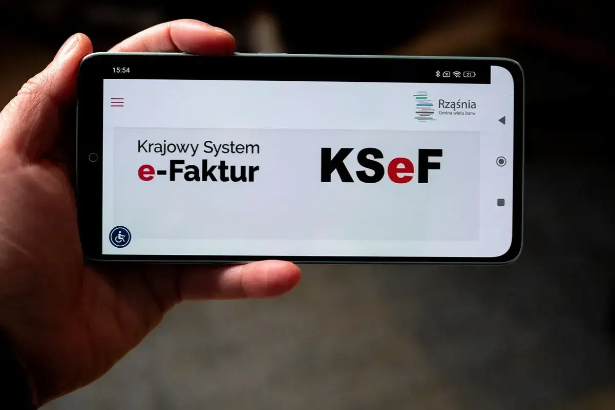 Dłoń trzyma smartfon z widocznym ekranem Krajowego Systemu e-Faktur. Widnieje logo KSeF, sugerujące problem z logowaniem.