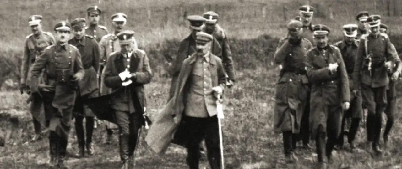 Grupa żołnierzy w mundurach maszeruje przez pole. Wśród nich widać postacie przypominające bolszewików, idących w zwartej grupie.