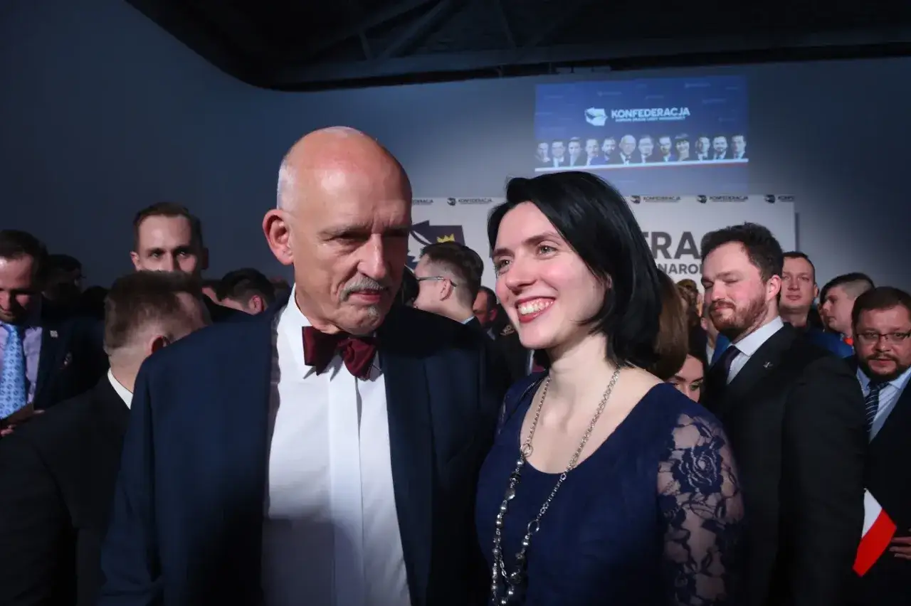 Janusz Korwin-Mikke z uśmiechniętą kobietą na tle Konfederacji. W tle widać innych mężczyzn, być może jego dzieci.