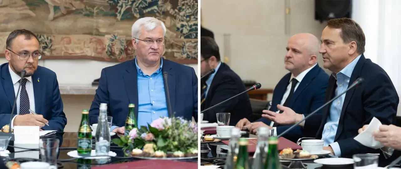 Dyplomata w garniturze z ukraińską flagą na klapie rozmawia przy stole z innymi mężczyznami.