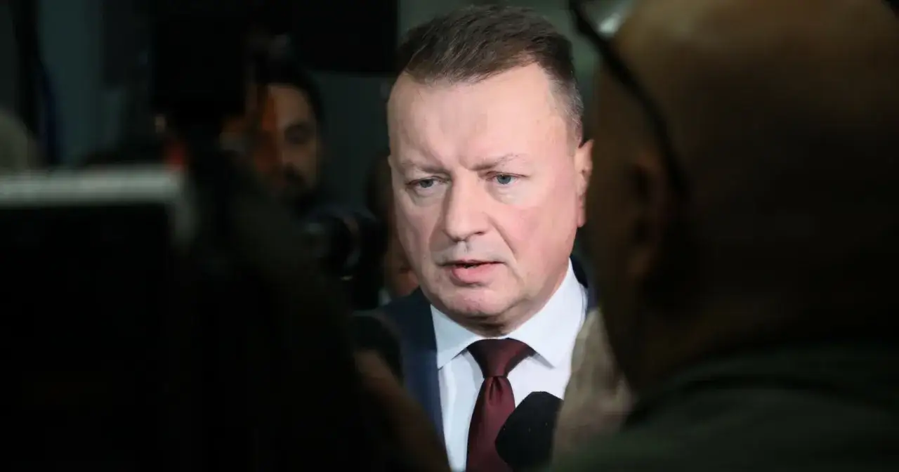 Mężczyzna w garniturze i krawacie, otoczony przez dziennikarzy. W tle widać inne osoby. Temat: zarzuty dla byłych ministrów PiS.