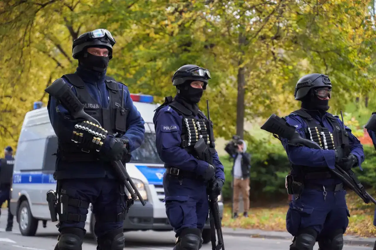 Trzech policjantów w pełnym rynsztunku, z bronią, czujnie obserwuje otoczenie. W tle radiowóz i drzewa. To obraz czujności i potencjalnej inwigilacji.