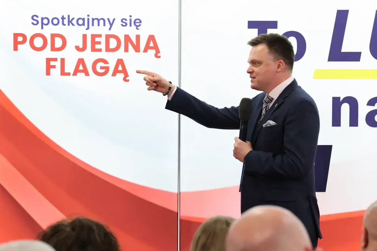 Mężczyzna w garniturze przemawia, wskazując na napis