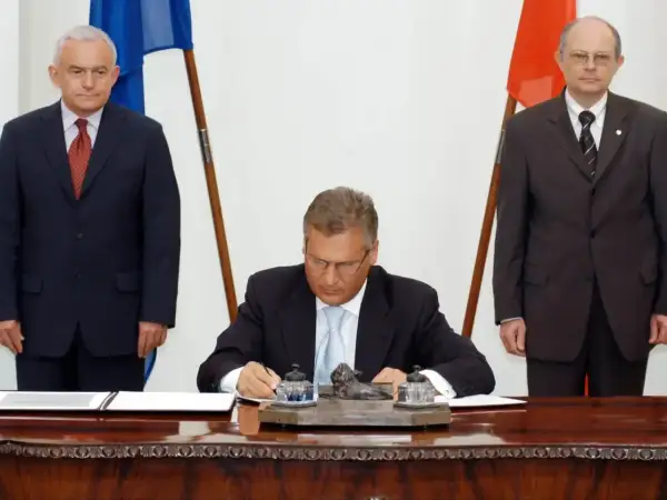 Uroczyste podpisanie dokumentów, symboliczny moment, kiedy Polska weszła do Unii. Trzech mężczyzn w garniturach, flagi Polski i UE.