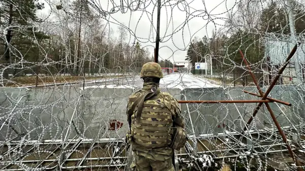 Żołnierz w mundurze obserwuje teren przez zasieki z drutu kolczastego. Widoczna jest granica polsko-białoruska, z budynkami i znakami po drugiej stronie.