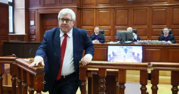 Mężczyzna w garniturze i krawacie wchodzi na salę sądową. Sędziowie w togach siedzą za stołem. Proces polityków PiS trwa.