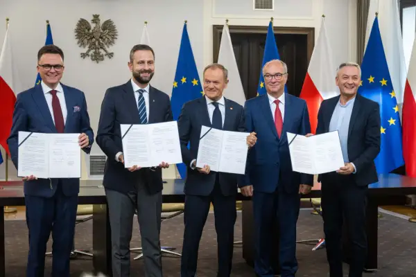 Pięciu mężczyzn w garniturach stoi przed flagami Polski i UE, trzymając dokumenty.