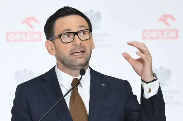 Mężczyzna w garniturze i okularach przemawia na tle logo Orlenu. Analiza fuzji Orlenu z Lotosem za rządów PiS.