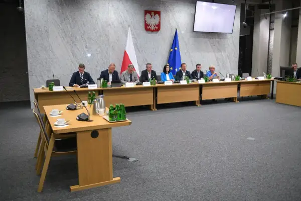 Osiem osób siedzi przy stole w sali konferencyjnej, przed nimi flagi Polski i UE.