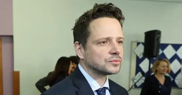Rafał Trzaskowski, którego wykształcenie budzi zainteresowanie, w garniturze i krawacie. W tle inne osoby i tablica.